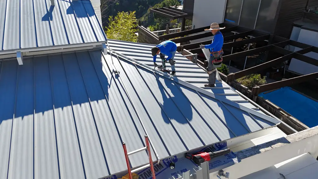 Two crew members working across a light gray standing seam roof beside a pool