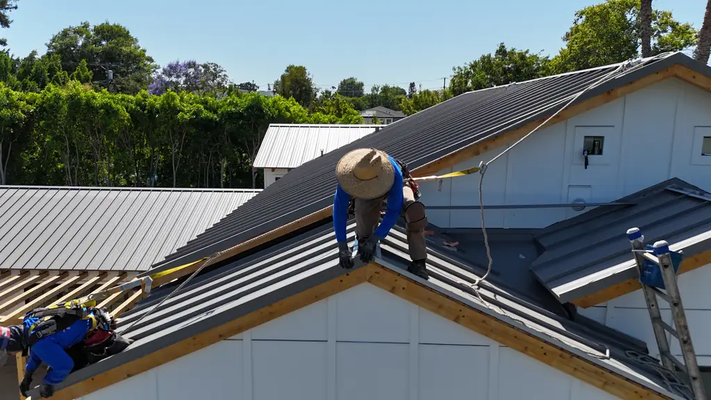 Crew member in a straw hat setting a dark standing seam panel at the ridge of a white board-and-batten house