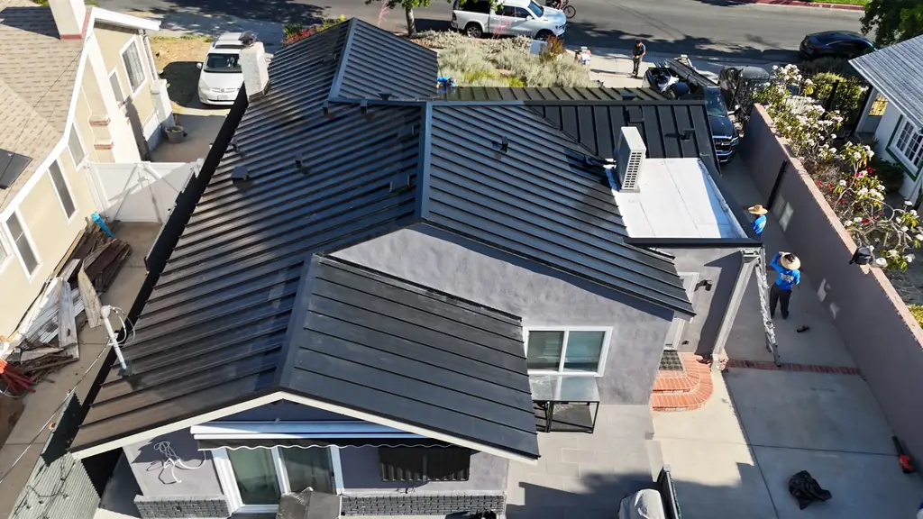 Aerial view of a new black standing seam roof on a stucco house, with a crew member on a ladder